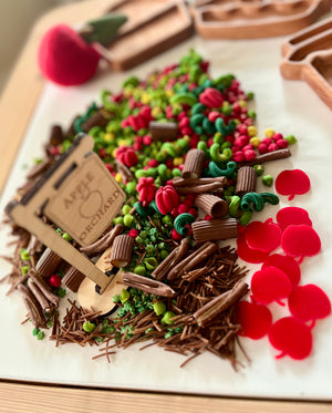 Miniature clay and felt play food pieces including red apples, green vegetables, and brown twigs arranged near a wooden sign on a white surface