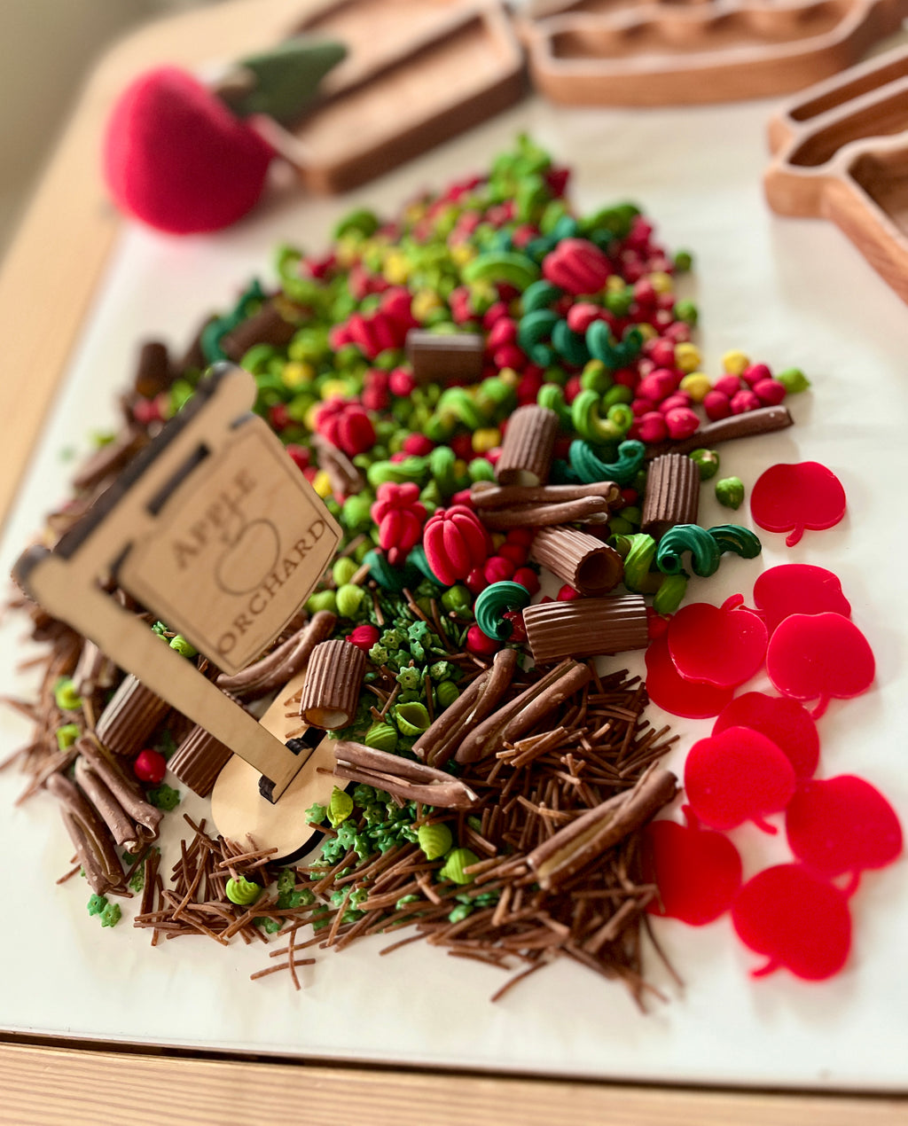 Miniature clay and felt play food pieces including red apples, green vegetables, and brown twigs arranged near a wooden sign on a white surface