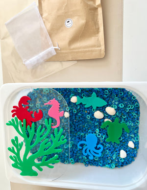 Plastic container with blue circular beads, white seashells, and colorful sea creature shapes on beige surface