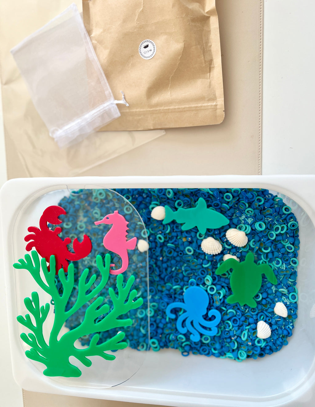 Plastic container with blue circular beads, white seashells, and colorful sea creature shapes on beige surface