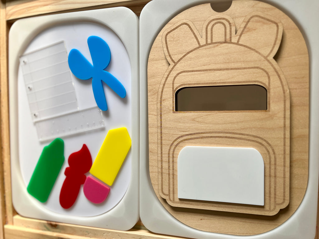 Wooden backpack-shaped puzzle with cutout school supplies and colorful translucent plastic pieces on white trays