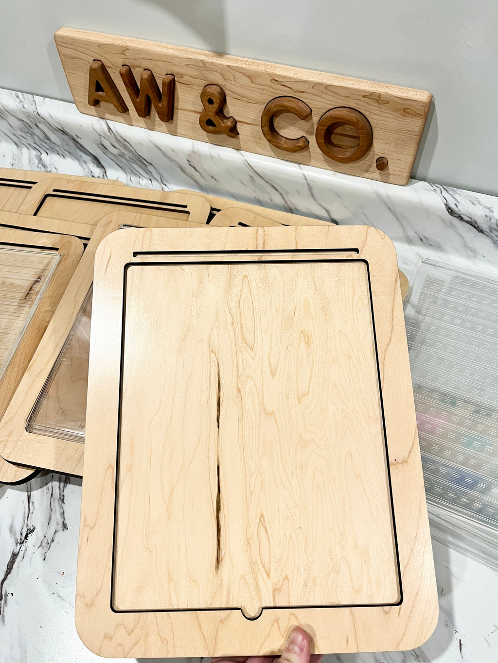 Light wooden tray with a rectangular groove held above other trays on a marble-patterned surface under a wooden AW & CO sign