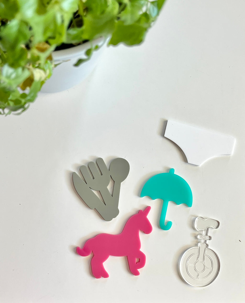 Assorted colorful acrylic shapes including gray cutlery, pink unicorn, teal umbrella, clear unicycle, and white arch on white surface with green plant above