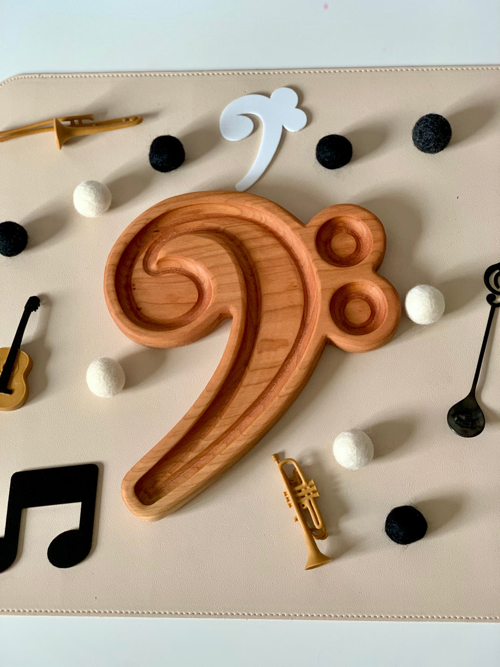 Wooden bass clef-shaped tray surrounded by black and white felt balls, miniature instruments, and music note cutouts on beige surface
