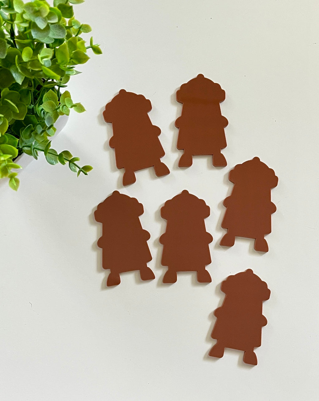 Six brown character-shaped cutouts arranged on a white surface near a green leafy plant