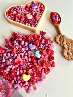 Assorted pink, red, and purple heart and flower-shaped pasta scattered next to a floral-carved wooden spoon and heart-shaped bowl