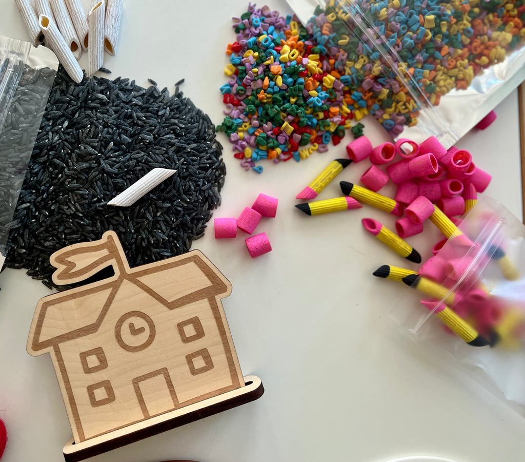 Wooden schoolhouse cutout with black rice, rainbow alphabet beads, pink erasers, and pencil-shaped craft pieces on white surface