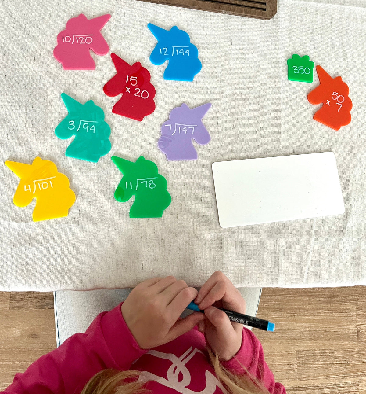 Colorful acrylic unicorn-shaped pieces with math problems written on them beside a blank white board, part of Little Dry Erasables - Set of 7 Large Unicorns, handcrafted homeschool education resources.