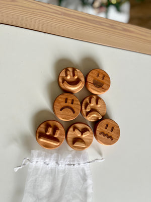 Seven round wooden tokens with carved expressive faces arranged on white surface near small sheer drawstring pouch