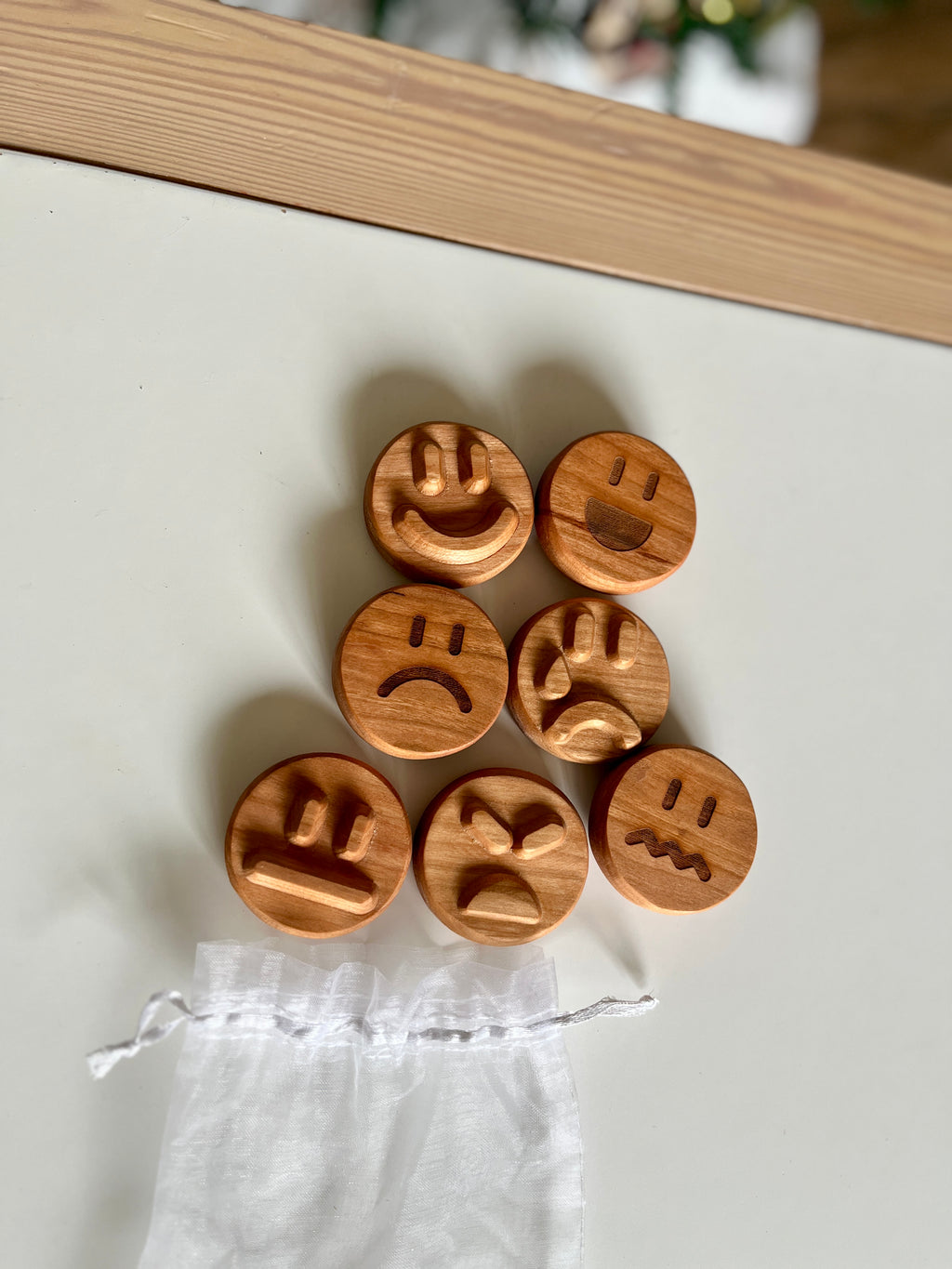 Seven round wooden tokens with carved expressive faces arranged on white surface near small sheer drawstring pouch
