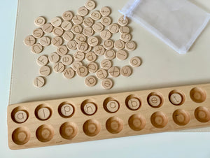 Small natural wooden alphabet discs with laser engraved letters scattered near storage pouch and wooden holder tray on light surface