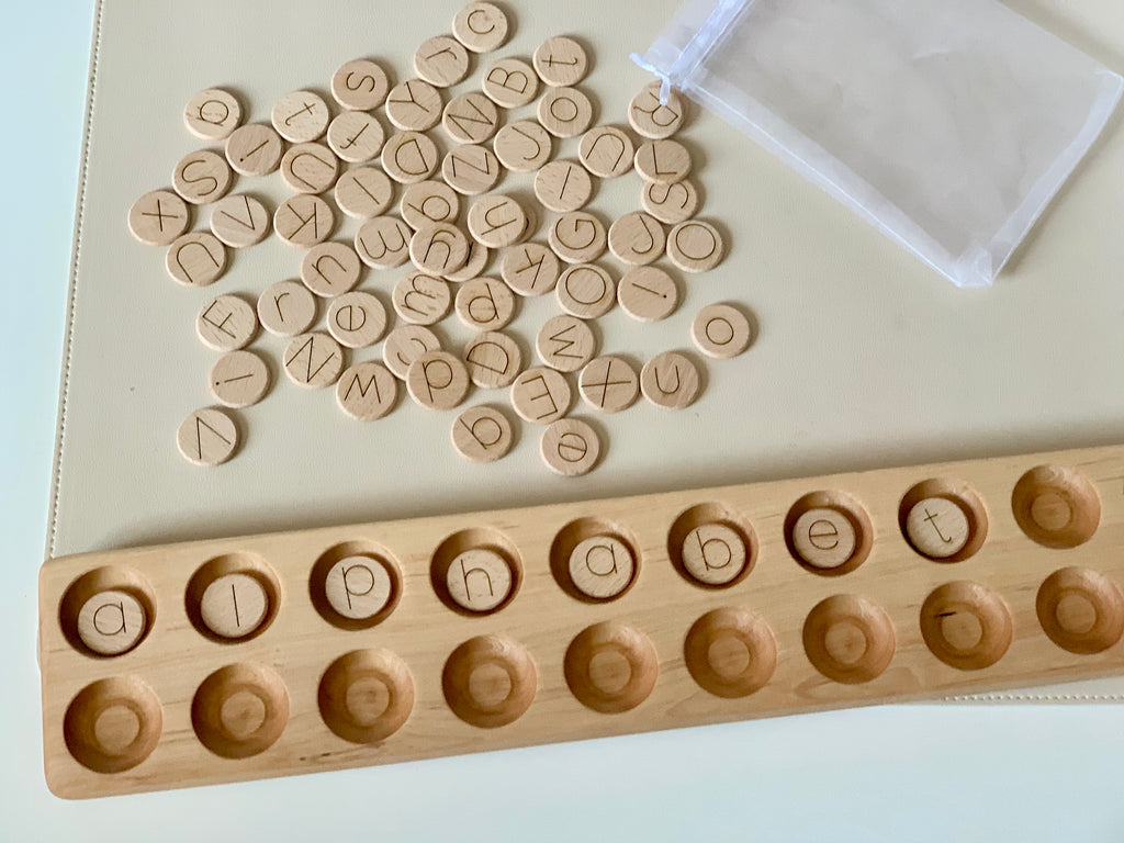 Small natural wooden alphabet discs with laser engraved letters scattered near storage pouch and wooden holder tray on light surface