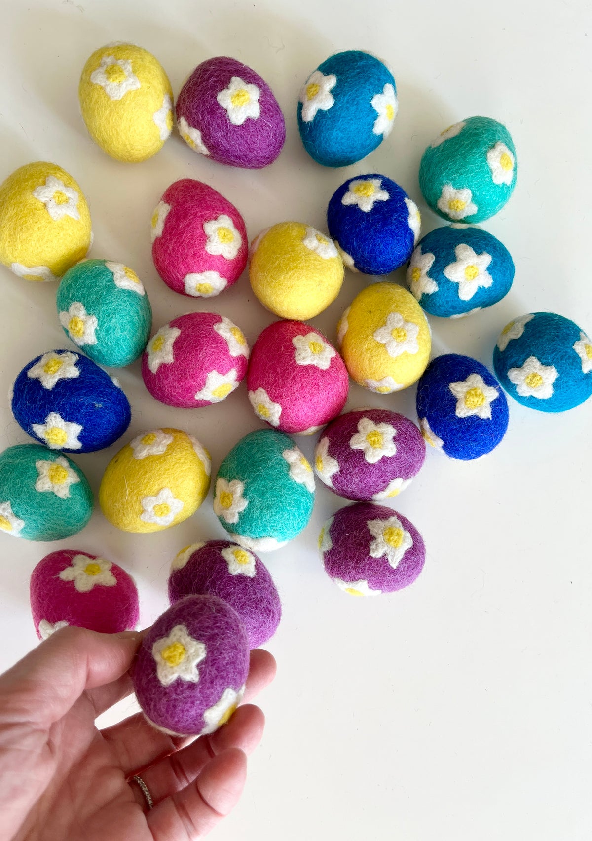 Hand holding purple felt egg with white flowers above scattered felt eggs in yellow, pink, blue, teal, and purple with floral designs on white surface