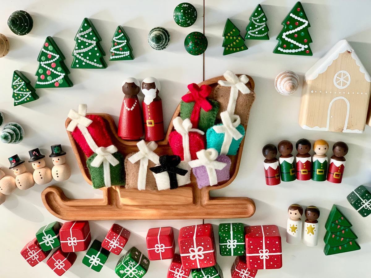 Hand-painted wooden Christmas peg dolls, gift boxes, trees, snowmen, and sleigh on white surface with gingerbread house