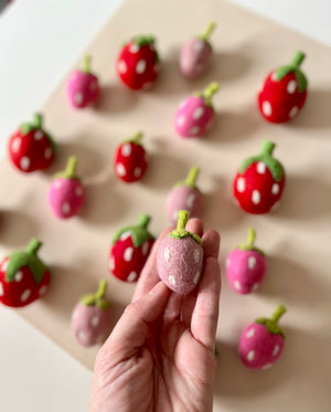 Hand holding a small pink felt strawberry with green stem and white seeds among scattered red and pink felt strawberries on beige background