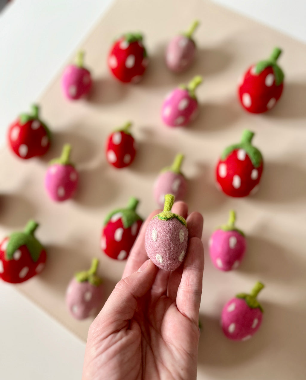 Hand holding a small pink felt strawberry with green stem and white seeds among scattered red and pink felt strawberries on beige background