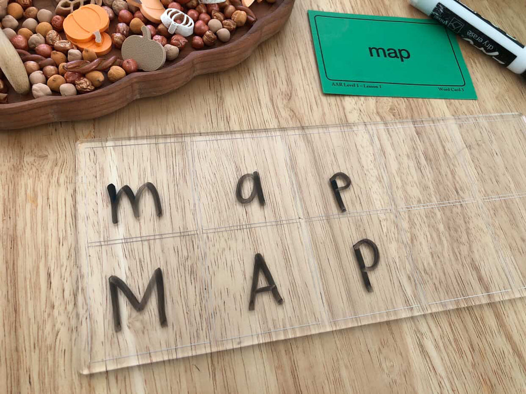 Transparent board with lowercase and uppercase letters m, a, p arranged in columns beside a green word card and dry erase marker