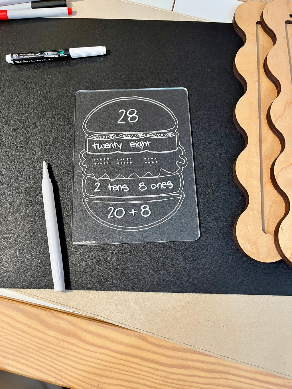 Clear plastic board with number 28 represented as digits, words, place value, and addition on a black surface with markers nearby