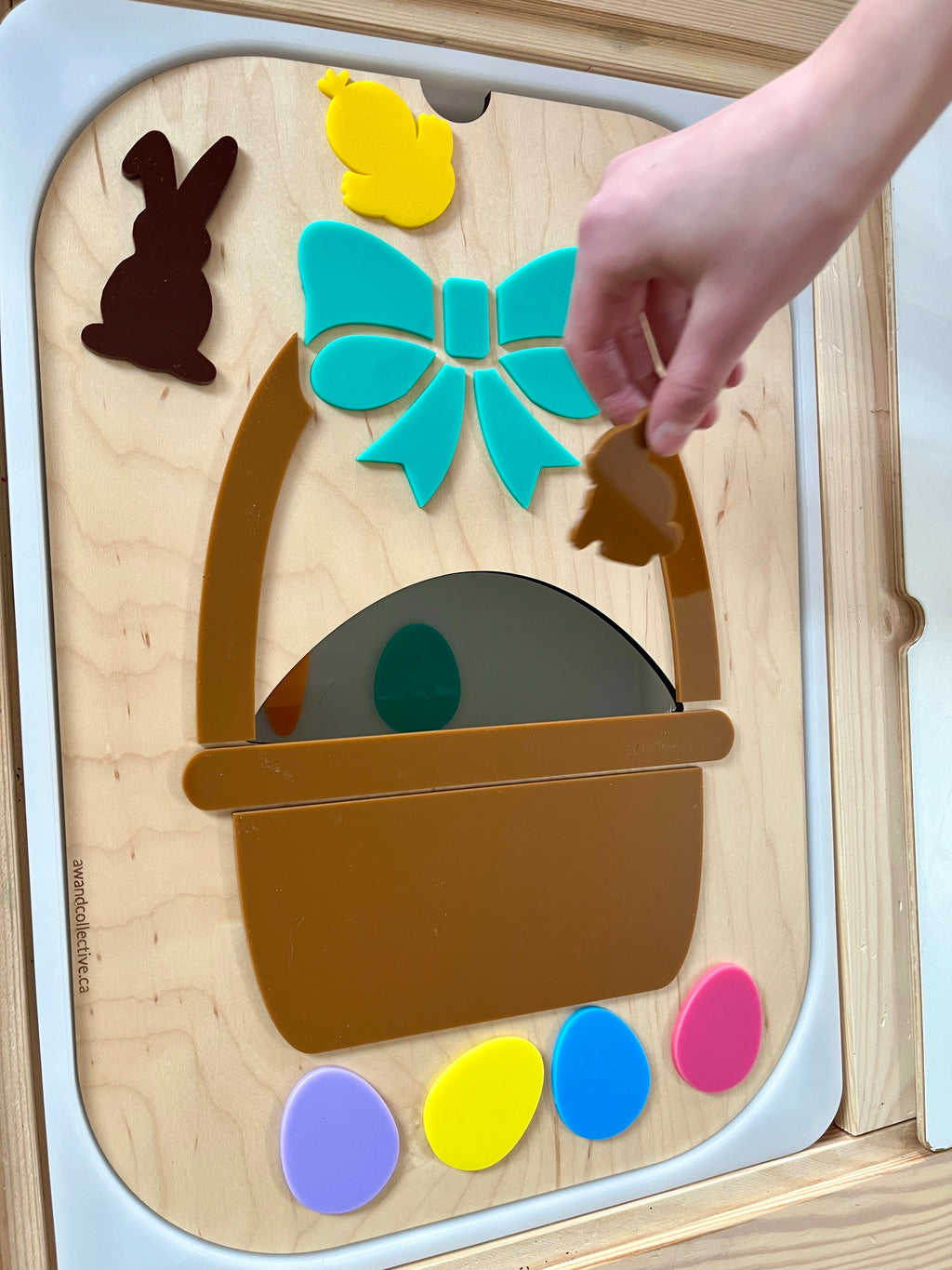 Child’s hand placing brown chick shape into a wooden board with a basket, bow, bunny, chick, and colorful eggs