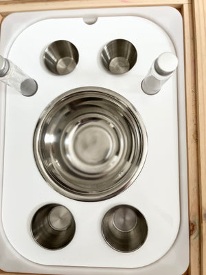 White tray holding stainless steel bowls and small glass vials on a wooden surface with rounded edges