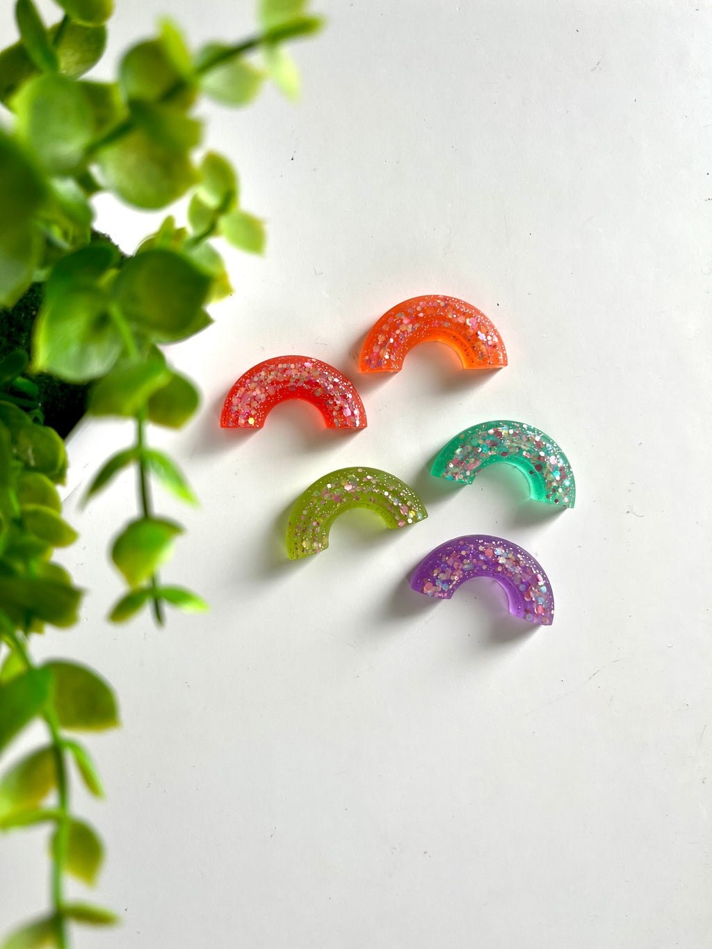 Five glitter-filled resin half-circle pieces in orange, red, green, teal, and purple on a white surface near green leaves