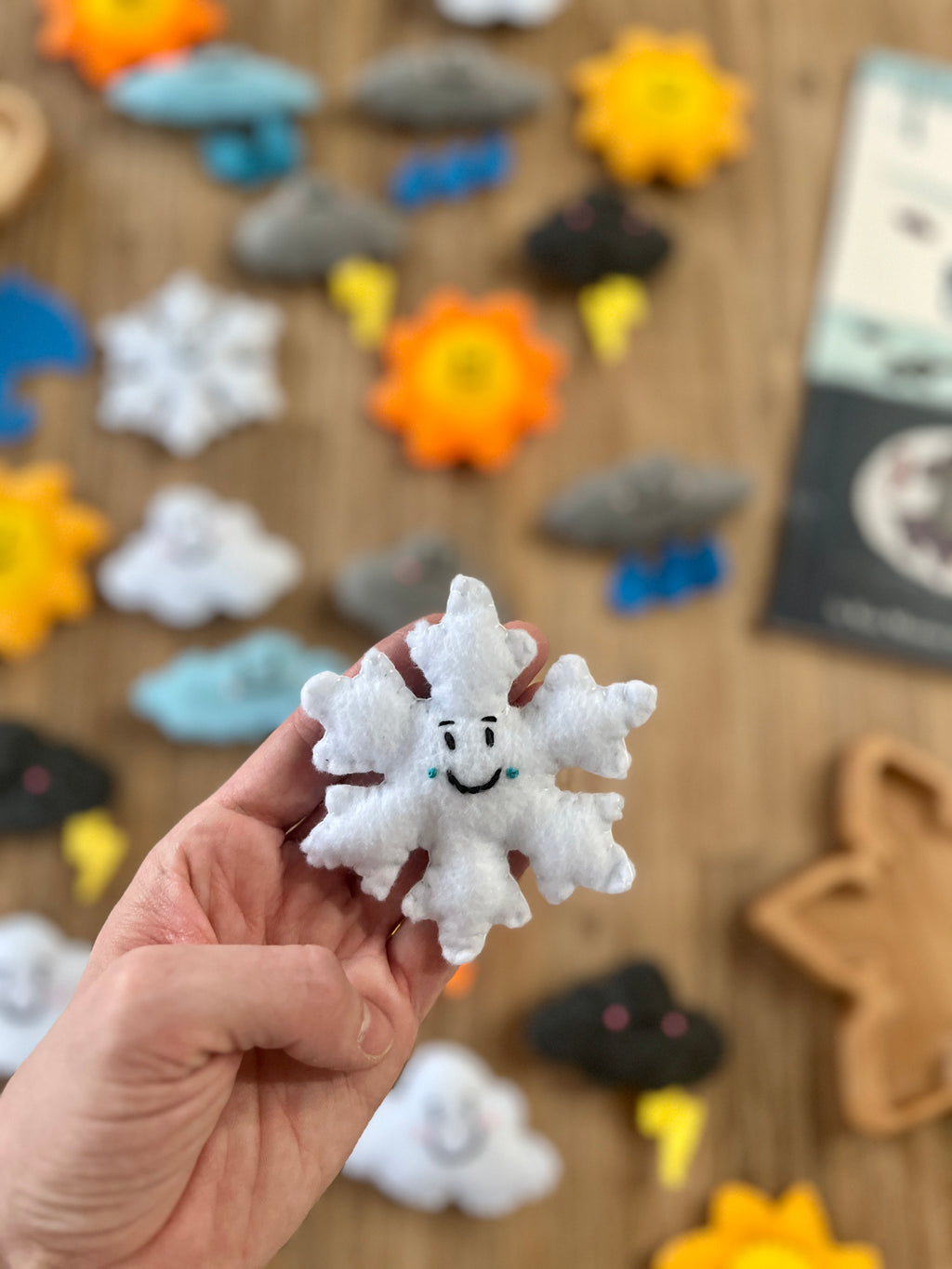 Hand holding a smiling white felt snowflake with blue teardrop details, surrounded by various colorful Felt Weather Shapes, Made in Canada, handcrafted, homeschool, education resources.