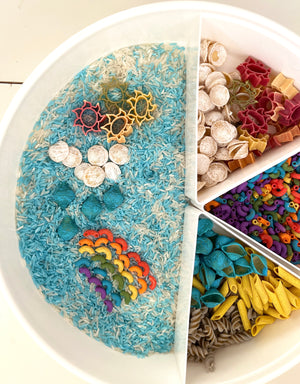 White divided tray with colorful pasta shapes, shells, and dyed rice arranged in rainbow, sea, and sky themes