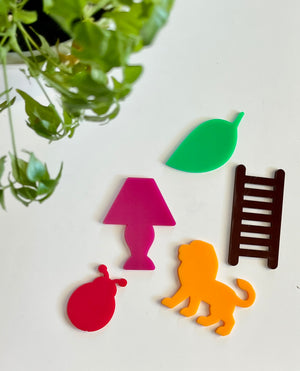 Five colorful acrylic shapes including pink ladybug, purple lamp, green leaf, brown ladder, and orange lion on white surface with green plant nearby