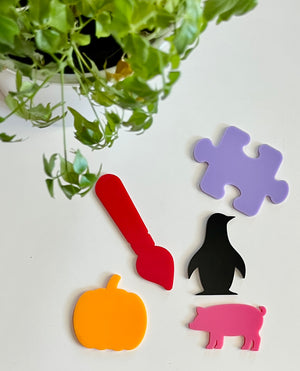 Five colorful acrylic shapes including orange pumpkin, red paintbrush, black penguin, pink pig, and purple puzzle piece on white surface with green leaves above