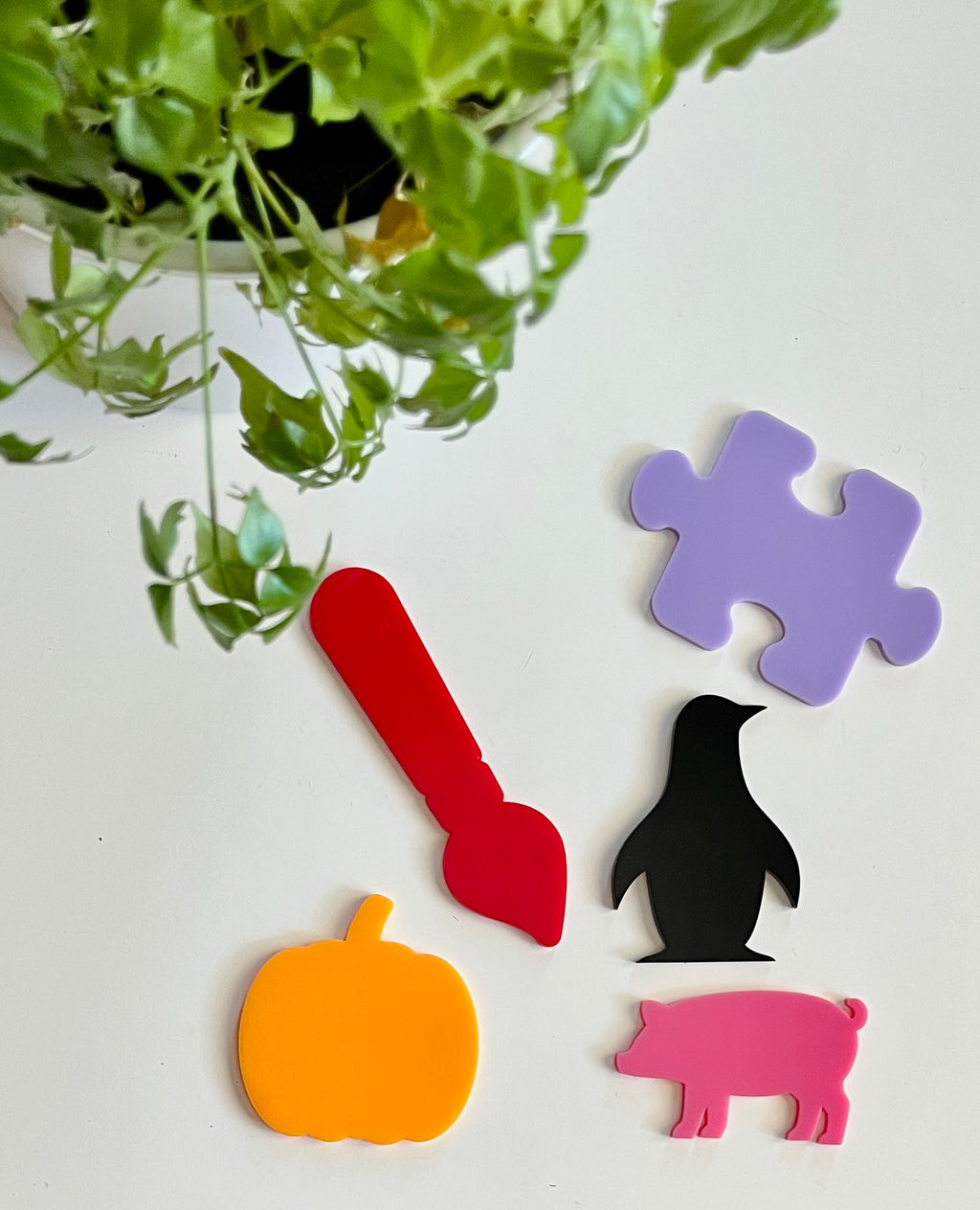 Five colorful acrylic shapes including orange pumpkin, red paintbrush, black penguin, pink pig, and purple puzzle piece on white surface with green leaves above