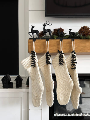 Four cream cable knit Christmas stockings hanging from wooden mantel with black cursive name tags and reindeer stocking holders