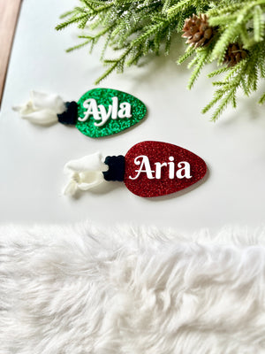 Two glittery Christmas light-shaped ornaments in red and green with white names and attached fabric tassels on white surface