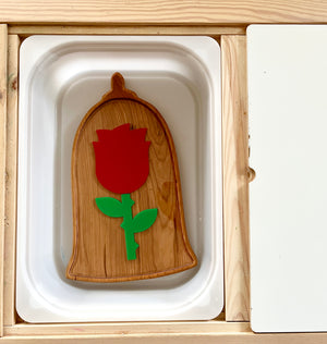 Wooden tray shaped like a bell with a red rose and green stem inside a white recessed frame on a wooden surface