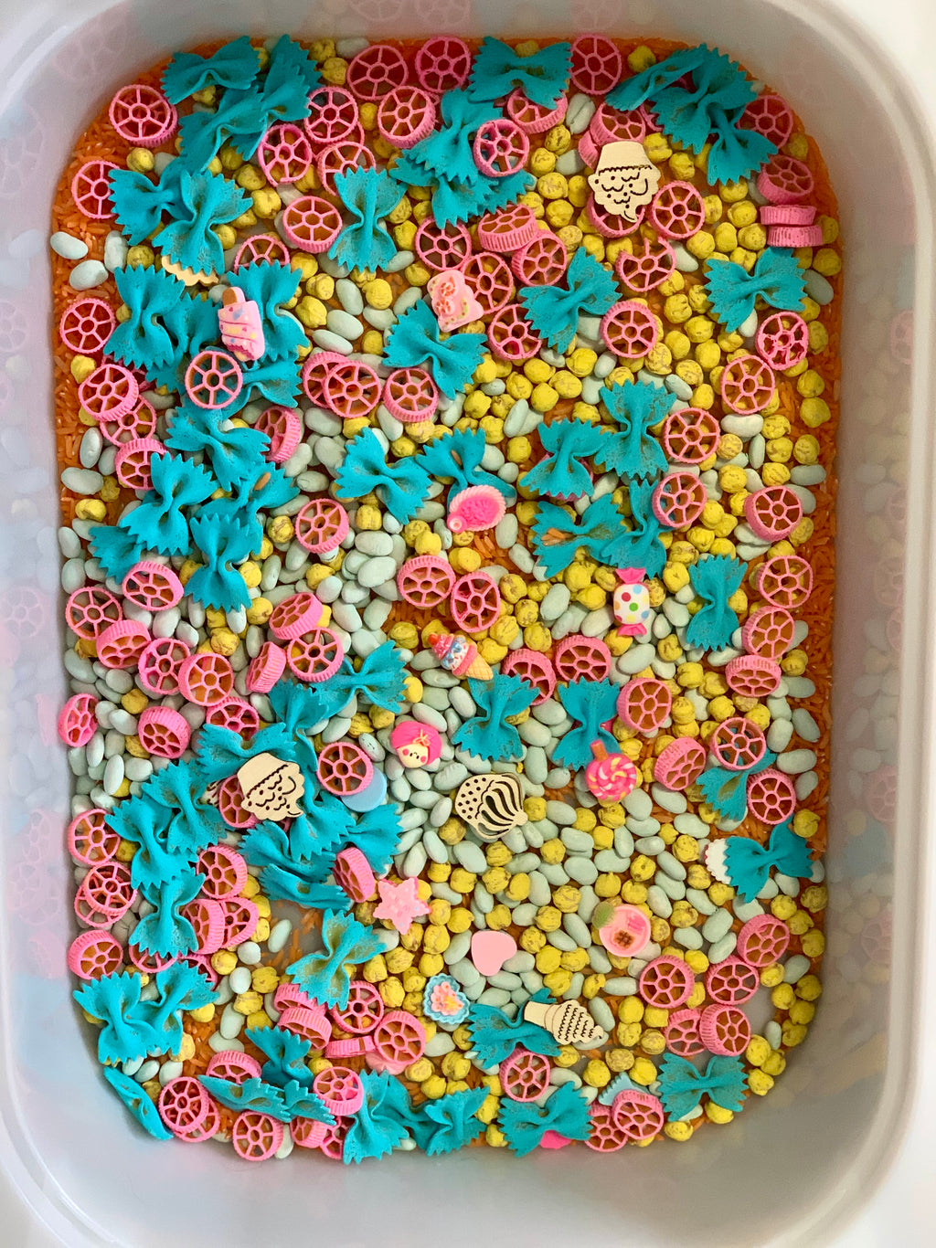 Plastic bin with yellow, white, blue bowtie pasta, pink wheel-shaped pasta, and small flat wooden shapes scattered inside