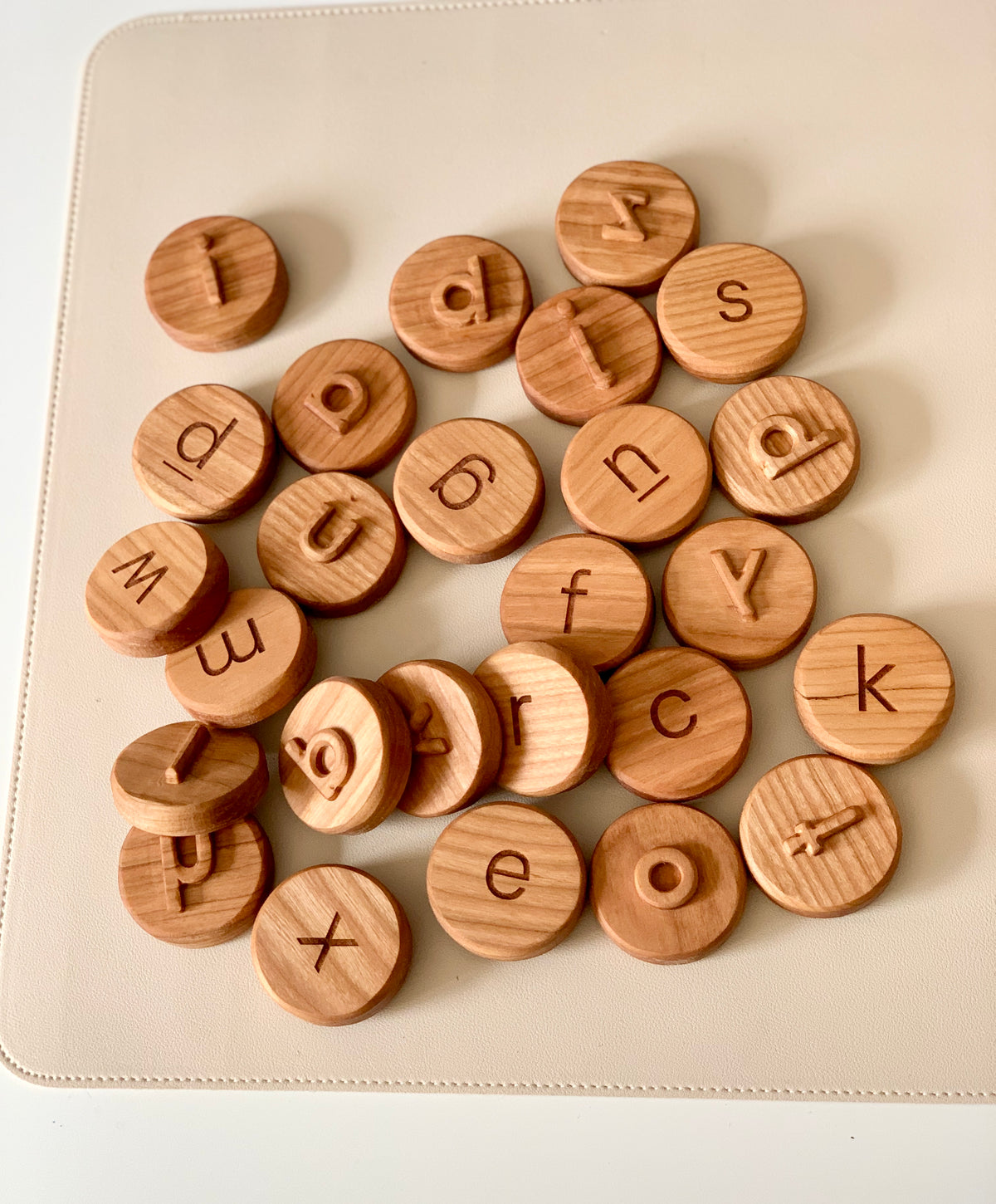 Scattered round wooden tokens engraved with lowercase and uppercase letters on a light beige surface