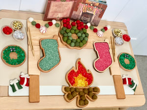 Wooden campfire sensory tray with green and red textured filling, felt garland, and holiday-themed decorations on light wood surface