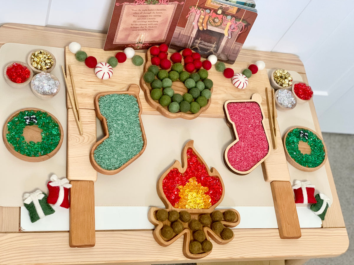 Wooden campfire sensory tray with green and red textured filling, felt garland, and holiday-themed decorations on light wood surface