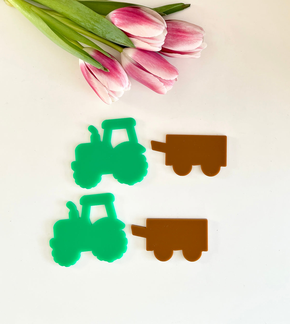 Two green tractor cutouts paired with two brown trailer cutouts beside a small bouquet of pink and white tulips on white surface