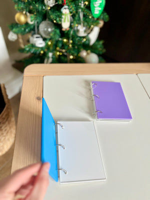 Two small ring binders with blank white and purple pages on a light wood and white table near a decorated Christmas tree