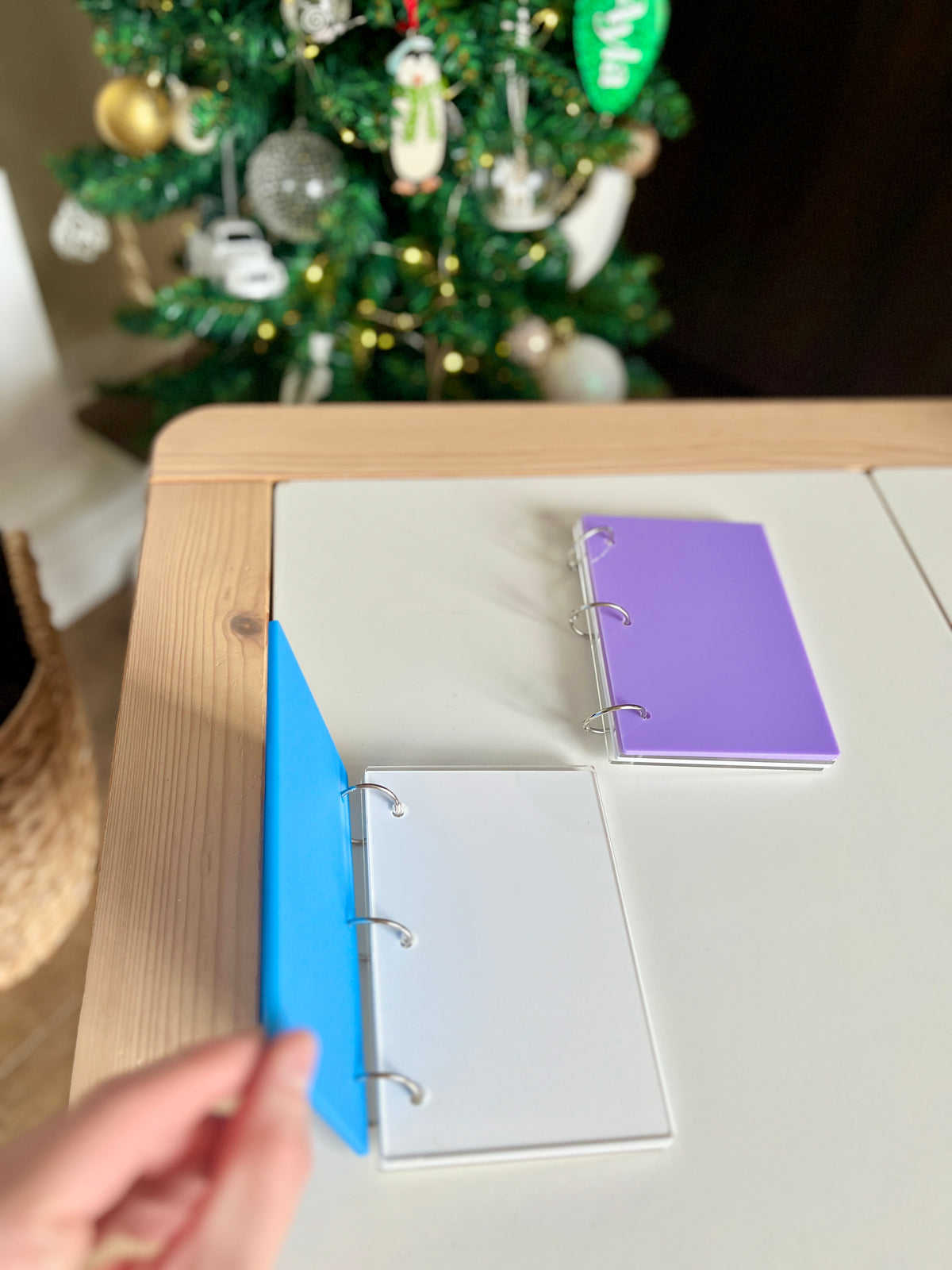 Two small ring binders with blank white and purple pages on a light wood and white table near a decorated Christmas tree