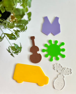 Five acrylic shapes including yellow van, brown violin, green virus, purple vest, and clear vase with flowers on white surface with green plant nearby