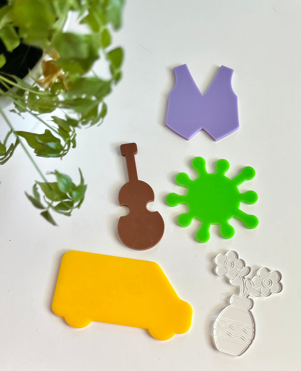Five acrylic shapes including yellow van, brown violin, green virus, purple vest, and clear vase with flowers on white surface with green plant nearby