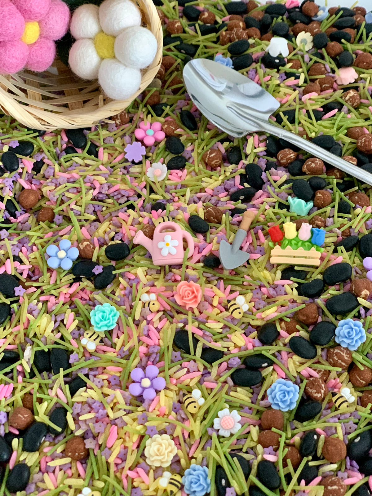 Colorful gardening-themed miniature toys and flower beads on a multicolored sensory bin with a small basket and spoon