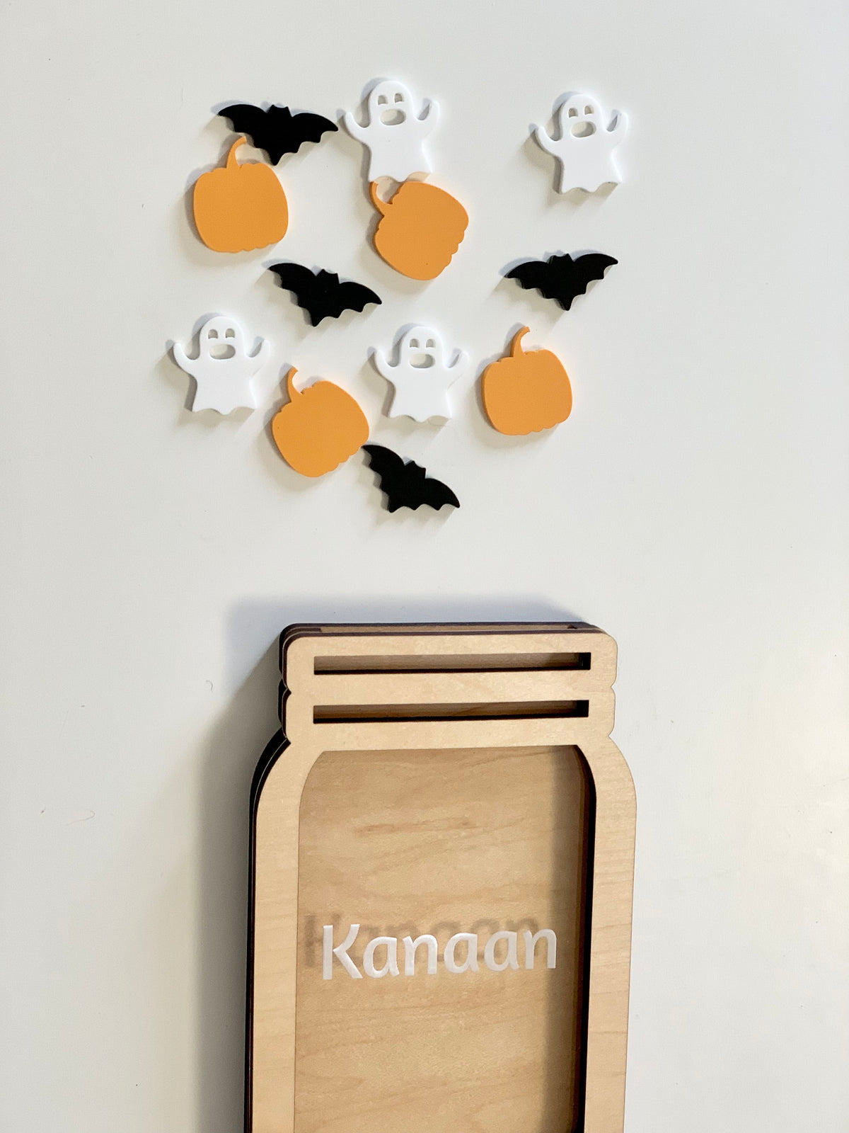 Light wooden jar-shaped frame with the name Kanaan and scattered orange pumpkin, white ghost, and black bat tokens above on a white background