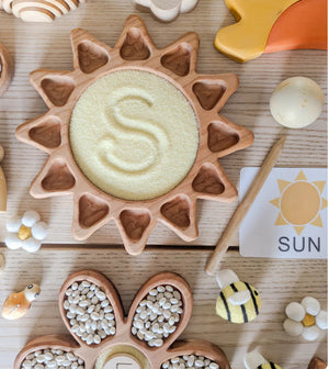 Wooden sun-shaped tray filled with yellow grains and surrounded by wooden toys and felt decorations on wood surface
