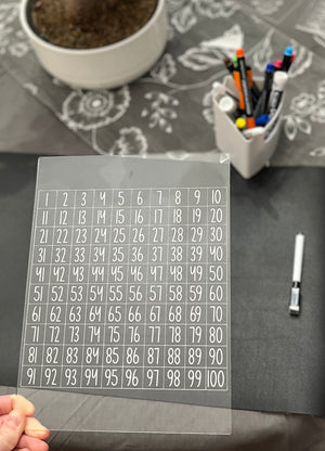 Clear acrylic sheet with a 1 to 100 numbered grid held above a dark surface with markers and a plant in background