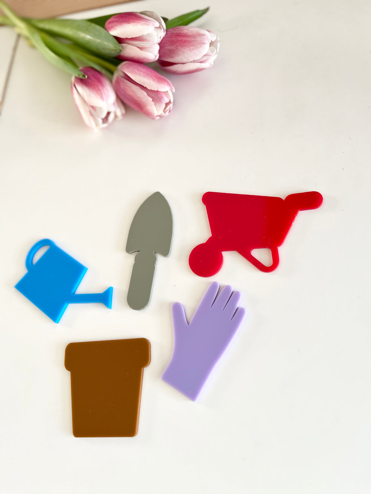 Pink tulips beside colorful flat gardening tool shapes including a blue watering can, gray trowel, red wheelbarrow, brown pot, and purple glove
