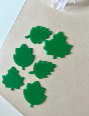 Six bright green plastic leaf-shaped cutouts of various designs arranged on a beige surface