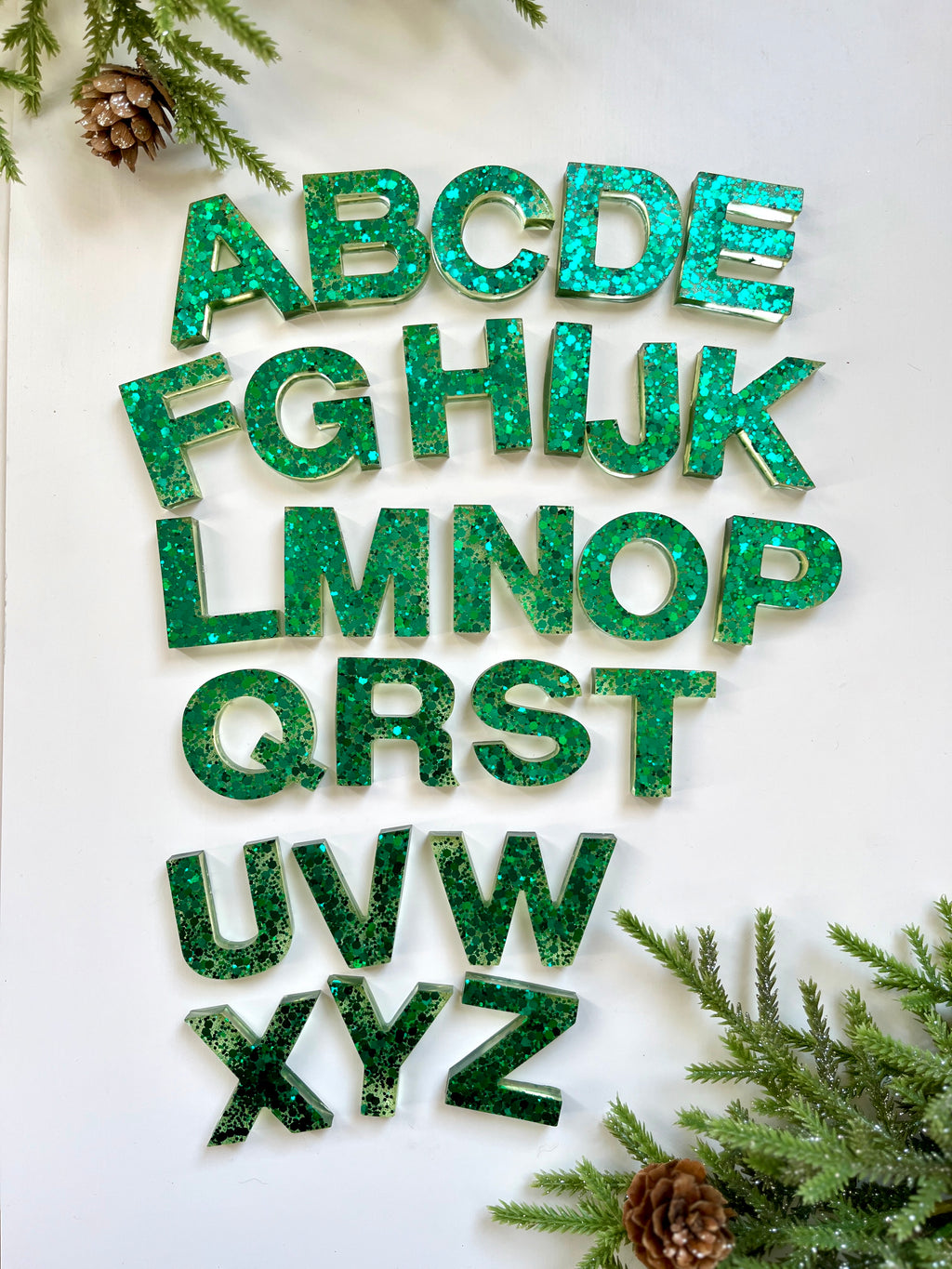 Green glitter resin alphabet letters arranged on white surface with pine cones and evergreen branches around edges