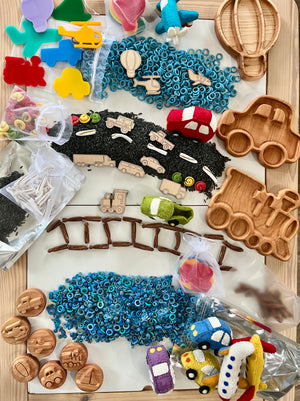 Wooden vehicle-shaped trays, colorful toy vehicles, and assorted sensory play materials arranged on a wooden surface.